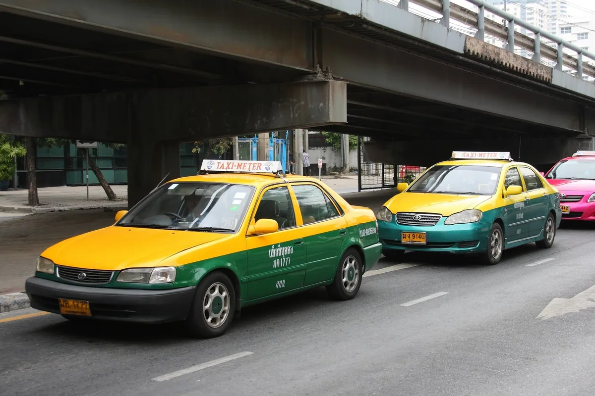 Two yellow-and-green Bangkok taxis with TAXI-METER roof signs waiting under an elevated road