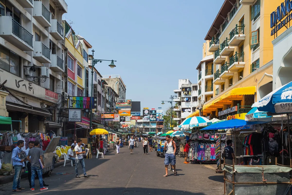 Khao San Road in Bangkok by day, lined with guesthouses, shop signs and market stalls under umbrellas