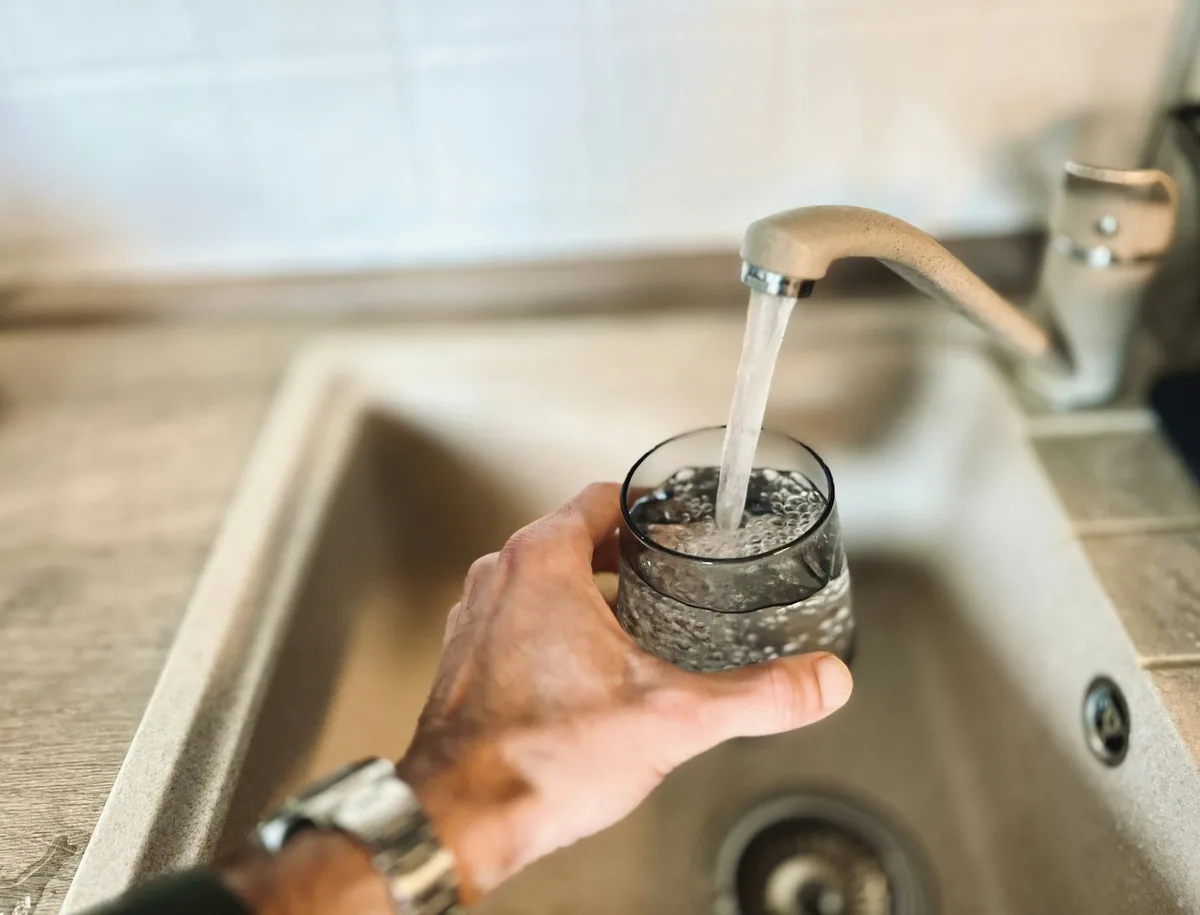 A hand filling a clear drinking glass with water straight from a kitchen tap over a sink