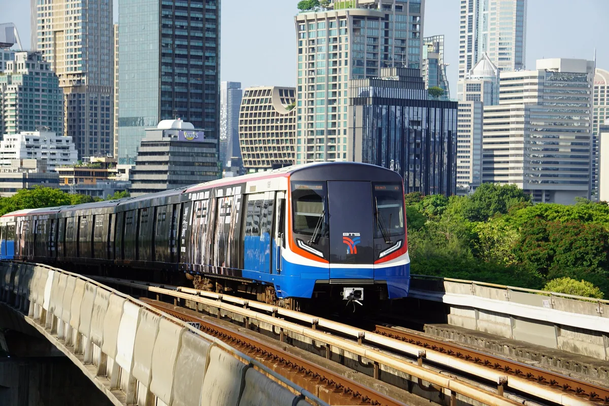 A blue-and-white BTS Skytrain on the elevated Silom Line track between Bangkok office towers