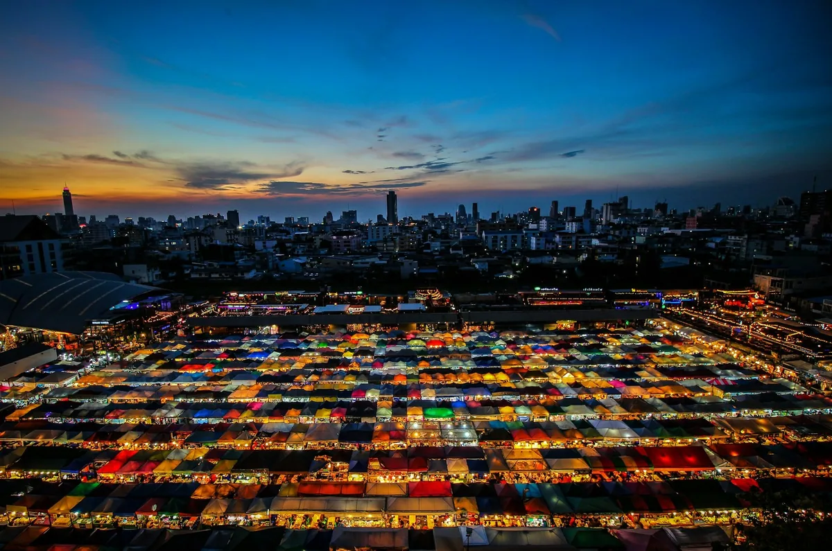 Rows of lit, multicoloured stall canopies at a Bangkok night market at dusk, with the city skyline behind