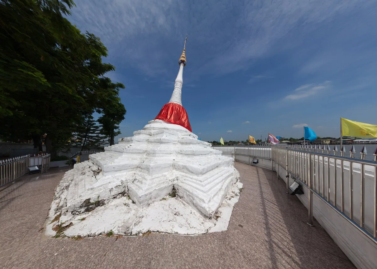 White Mon-style chedi wrapped in red cloth on the riverside walkway at Wat Paramai Yikawat, Ko Kret, with prayer flags beyond