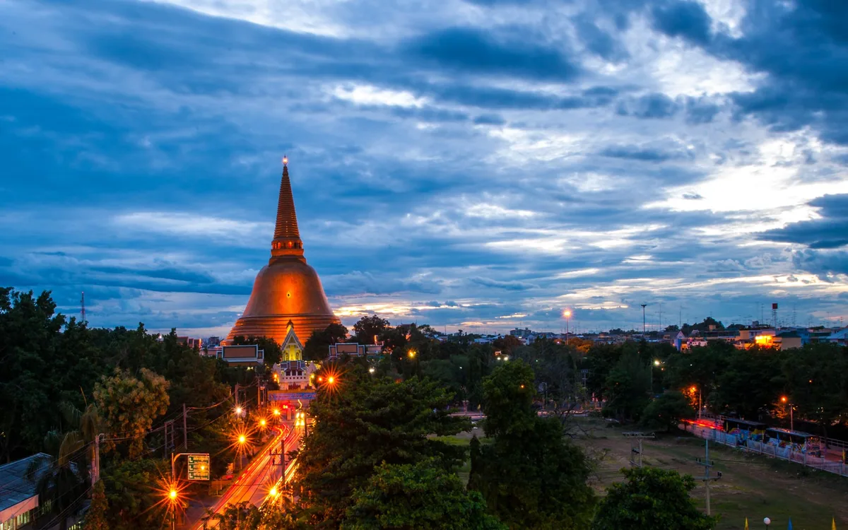 Floodlit Phra Pathom Chedi rising above the trees of Nakhon Pathom under a cloudy evening sky