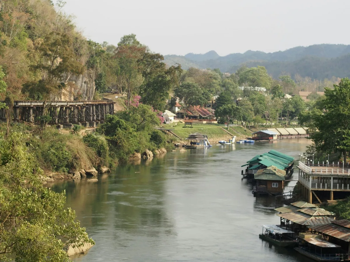 Wooden Death Railway viaduct along a cliff above the River Kwai with houseboats moored below