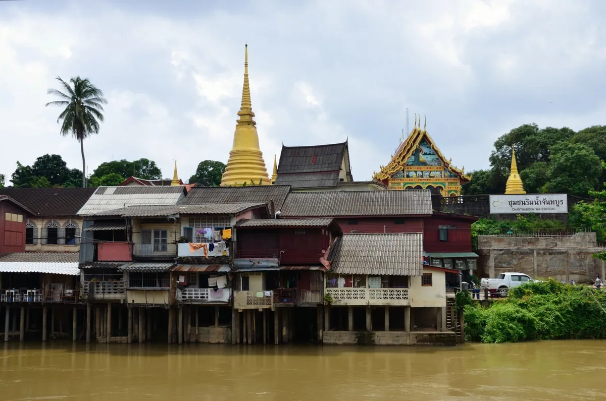 Old stilt houses of the Chanthaboon Waterfront community on the Chanthaburi River beneath a golden temple chedi