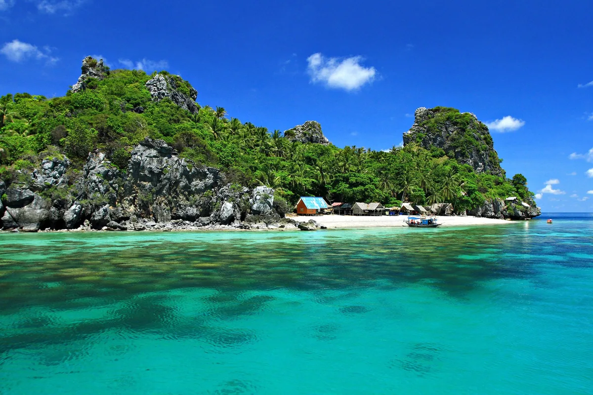 Turquoise water off a small beach with huts beneath limestone crags on an island in Mu Ko Chumphon National Park