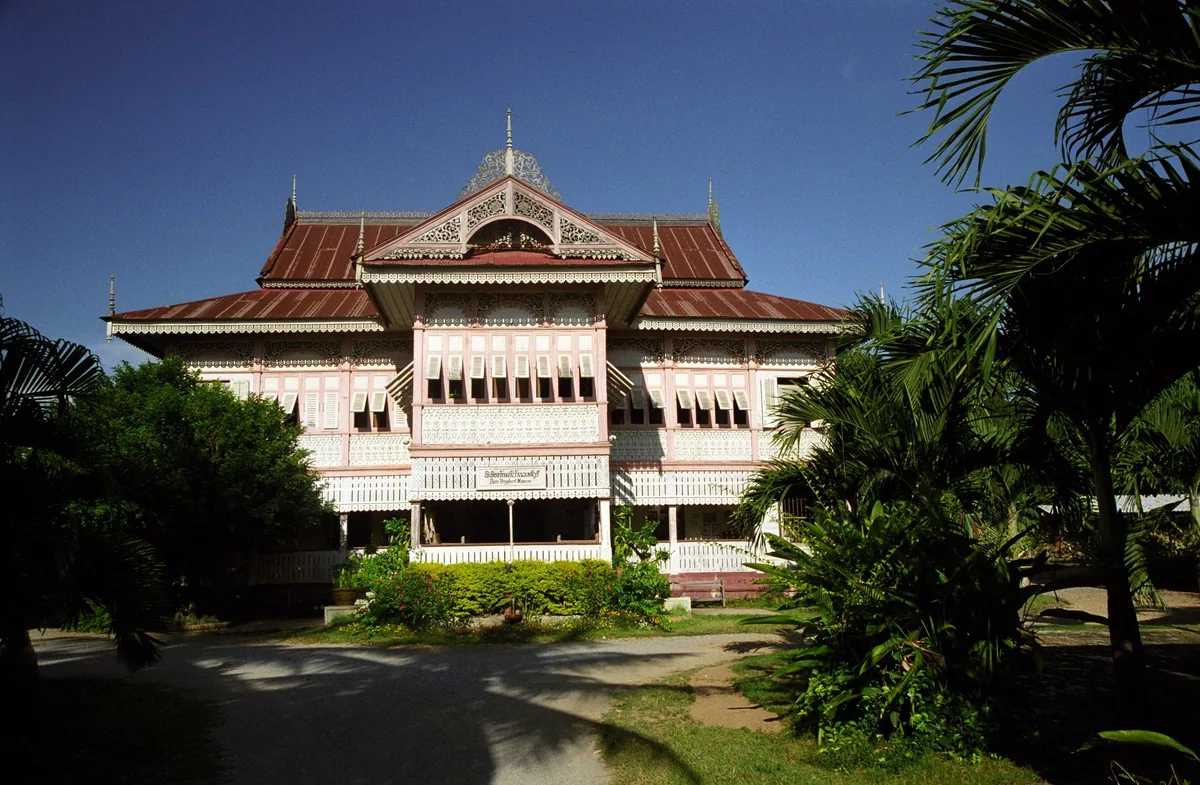 The pink-and-white teak mansion of Vongburi House in Phrae with carved fretwork eaves under a deep blue sky