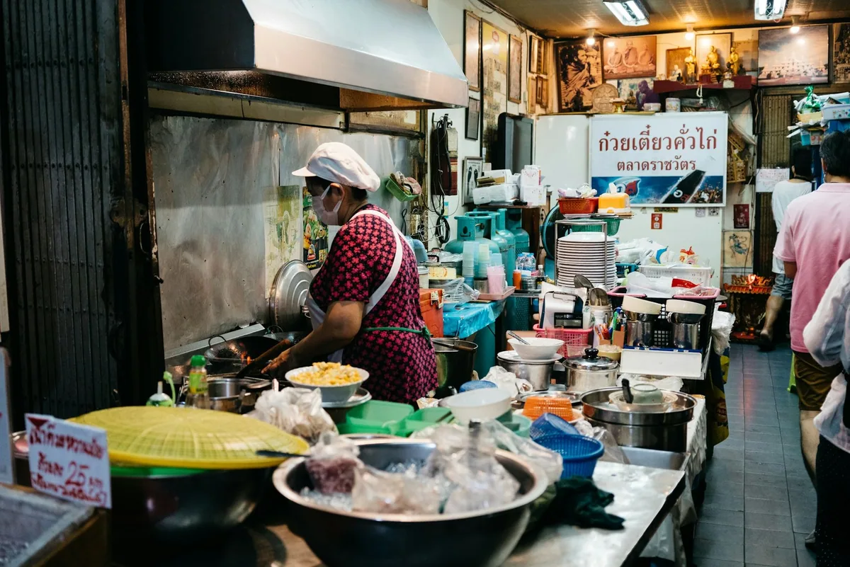 Cook in a mask frying rice noodles at a wok station in a crowded shophouse kitchen in Bangkok