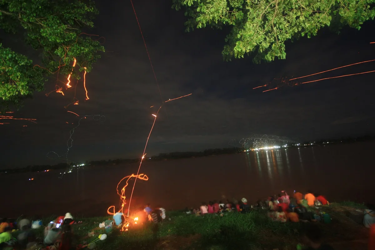 Crowds on the Mekong bank at night watching glowing streaks rise over the river at the end of Buddhist Lent