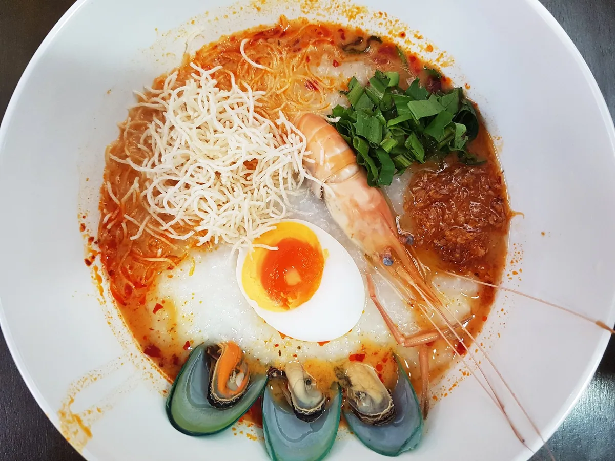Bowl of tom yam rice congee in Bangkok topped with a prawn, mussels, a soft-boiled egg and crispy noodles