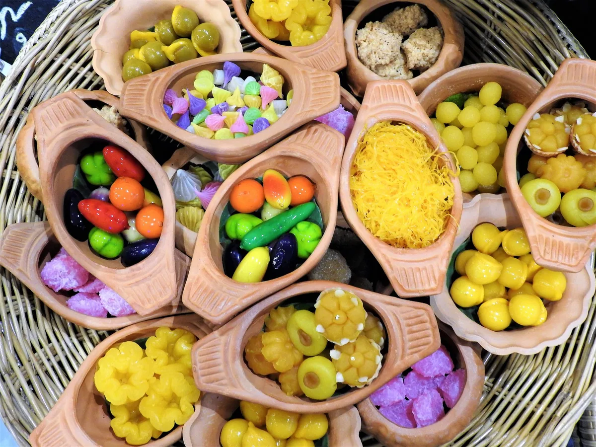 Clay dishes of traditional Thai sweets, including fruit-shaped luk chup and golden foi thong threads, in a woven tray