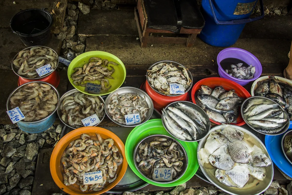 Bowls of fresh prawns, crabs and fish with handwritten Thai price tags laid out on the ground at a fresh market