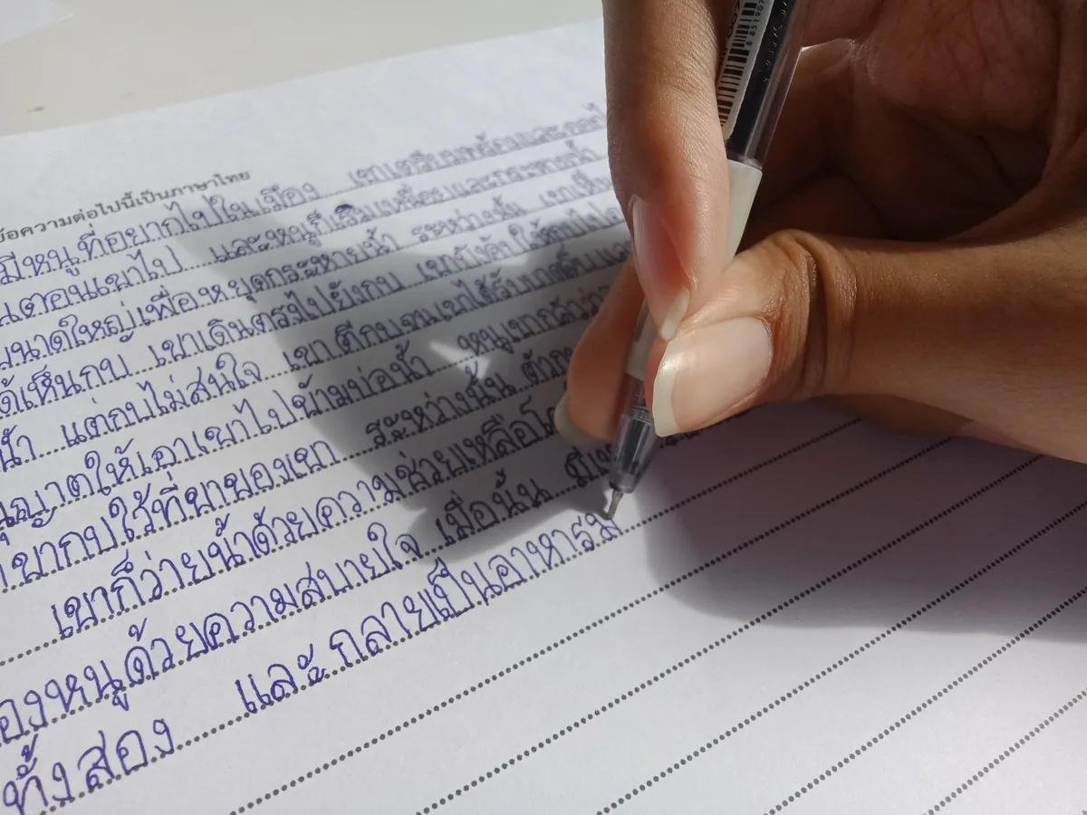 A hand writing lines of Thai script in blue ink with a ballpoint pen on ruled notebook paper