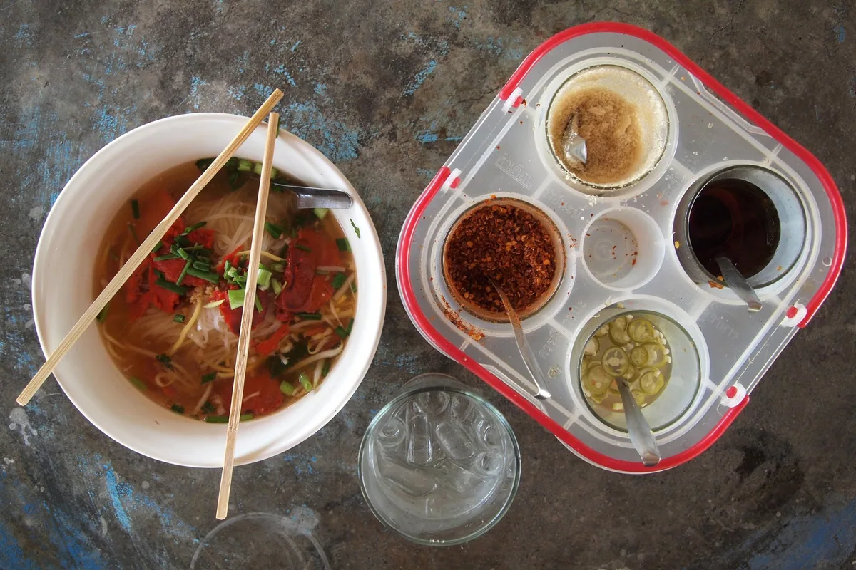 Bowl of rice noodle soup beside a condiment caddy with sugar, chilli flakes, fish sauce and chillies in vinegar