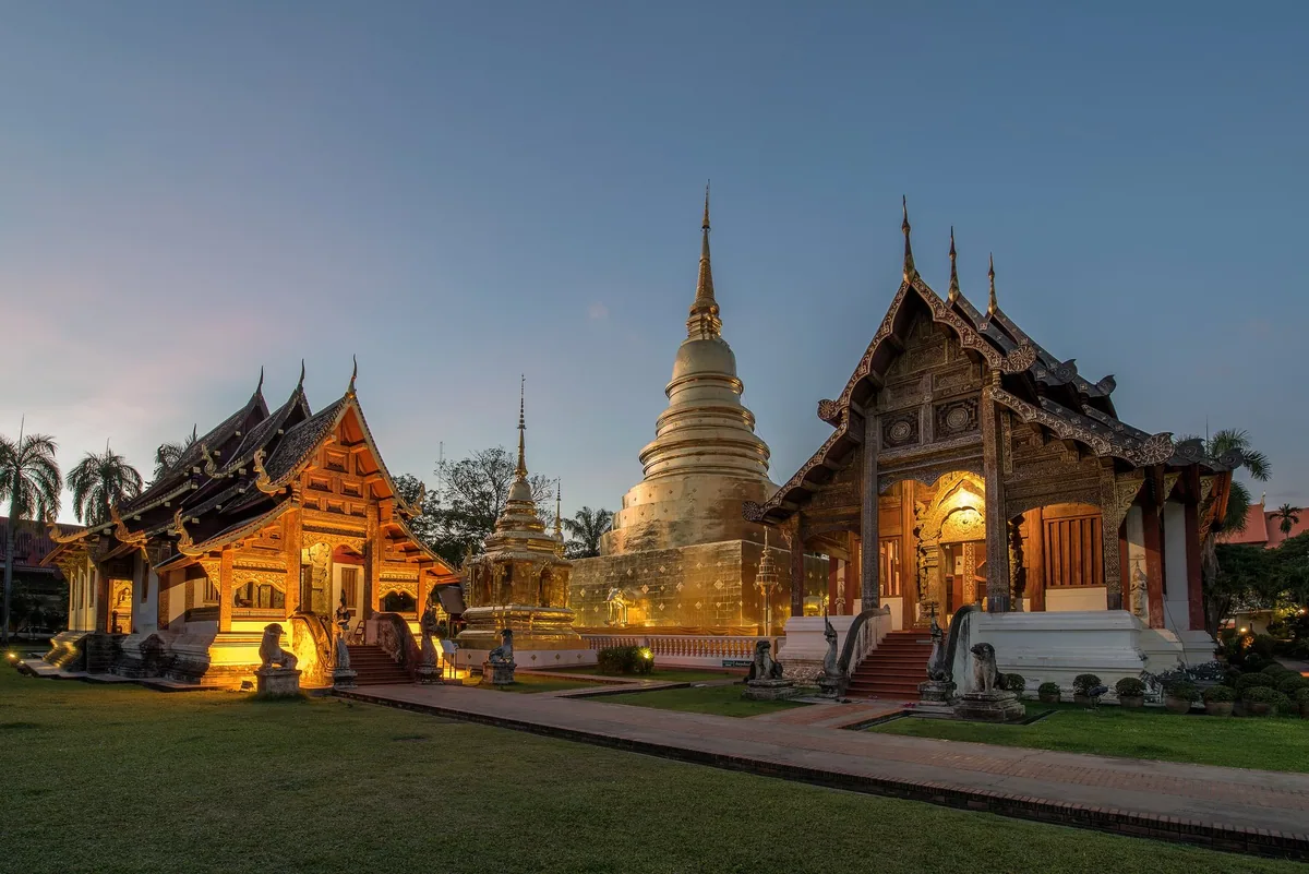 Two Lanna-style halls with tiered roofs flanking the gilded chedi of Wat Phra Singh in Chiang Mai at dusk