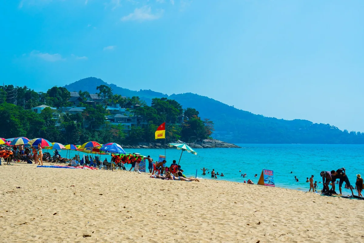 Sunbathers and red-and-yellow lifeguard flags on Surin Beach in Phuket under a clear sky