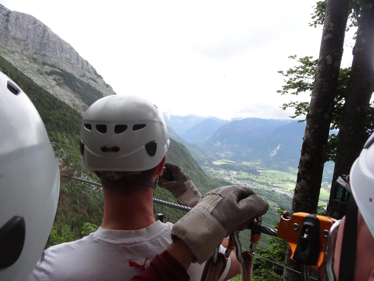 Helmeted riders in leather gloves clipped onto a zip-line cable with a pulley, looking out over a mountain valley