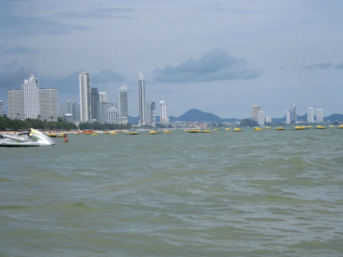 Choppy green seawater off Jomtien Beach, with a line of swimming-zone buoys, a jet ski and high-rise towers on the shore