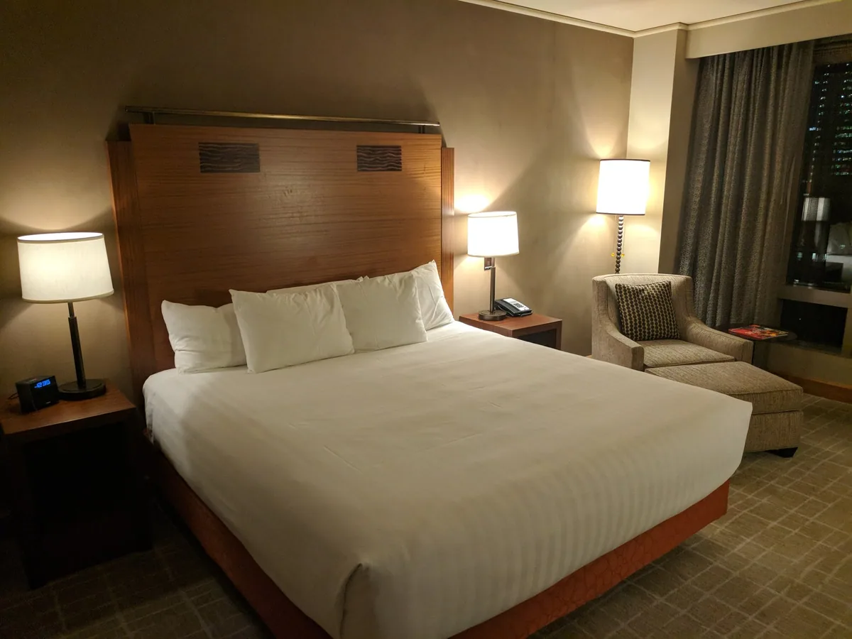 Neatly made hotel bed with white linen, a wooden headboard and bedside lamps in a dimly lit room