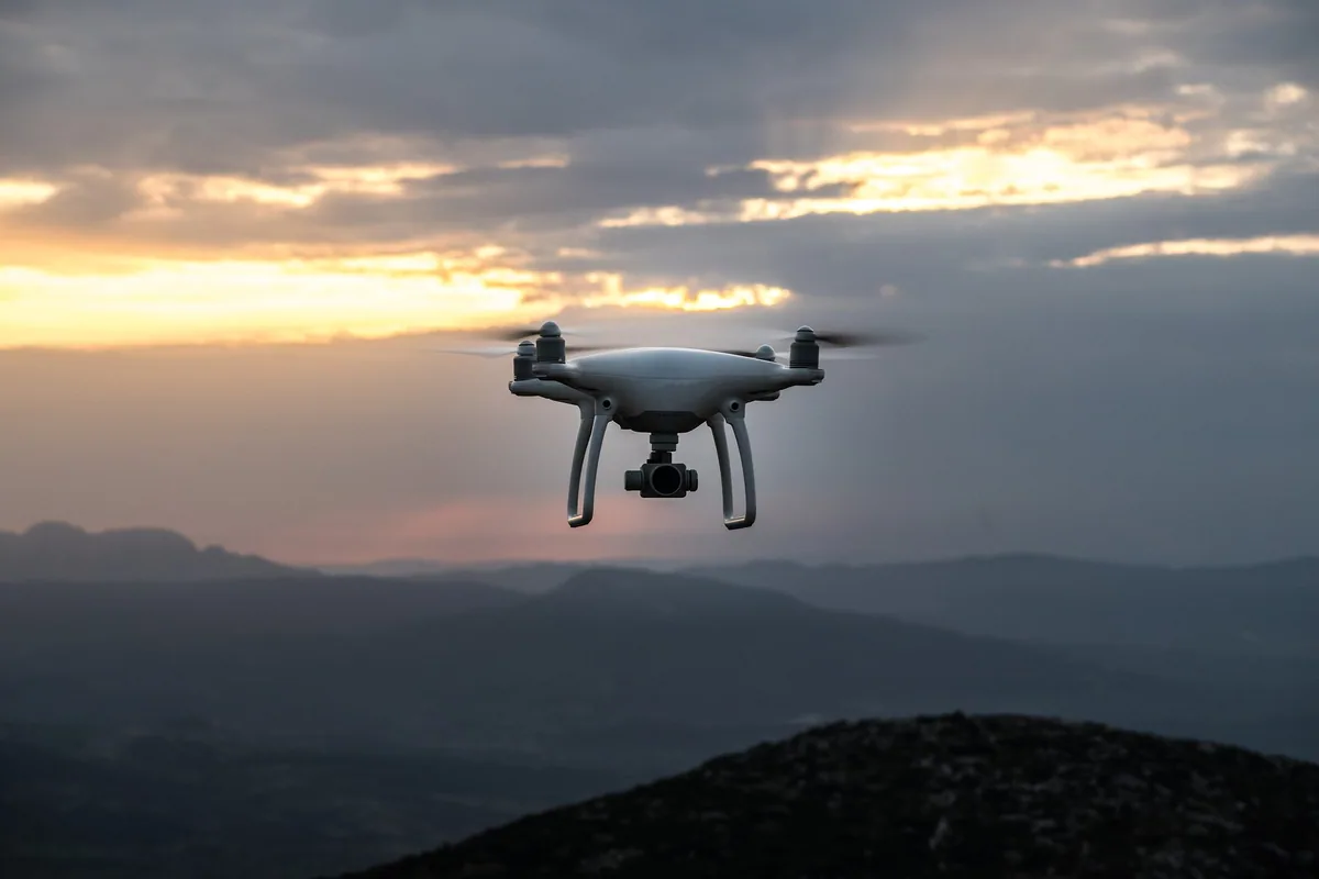 A white camera drone hovering against a cloudy evening sky above hills