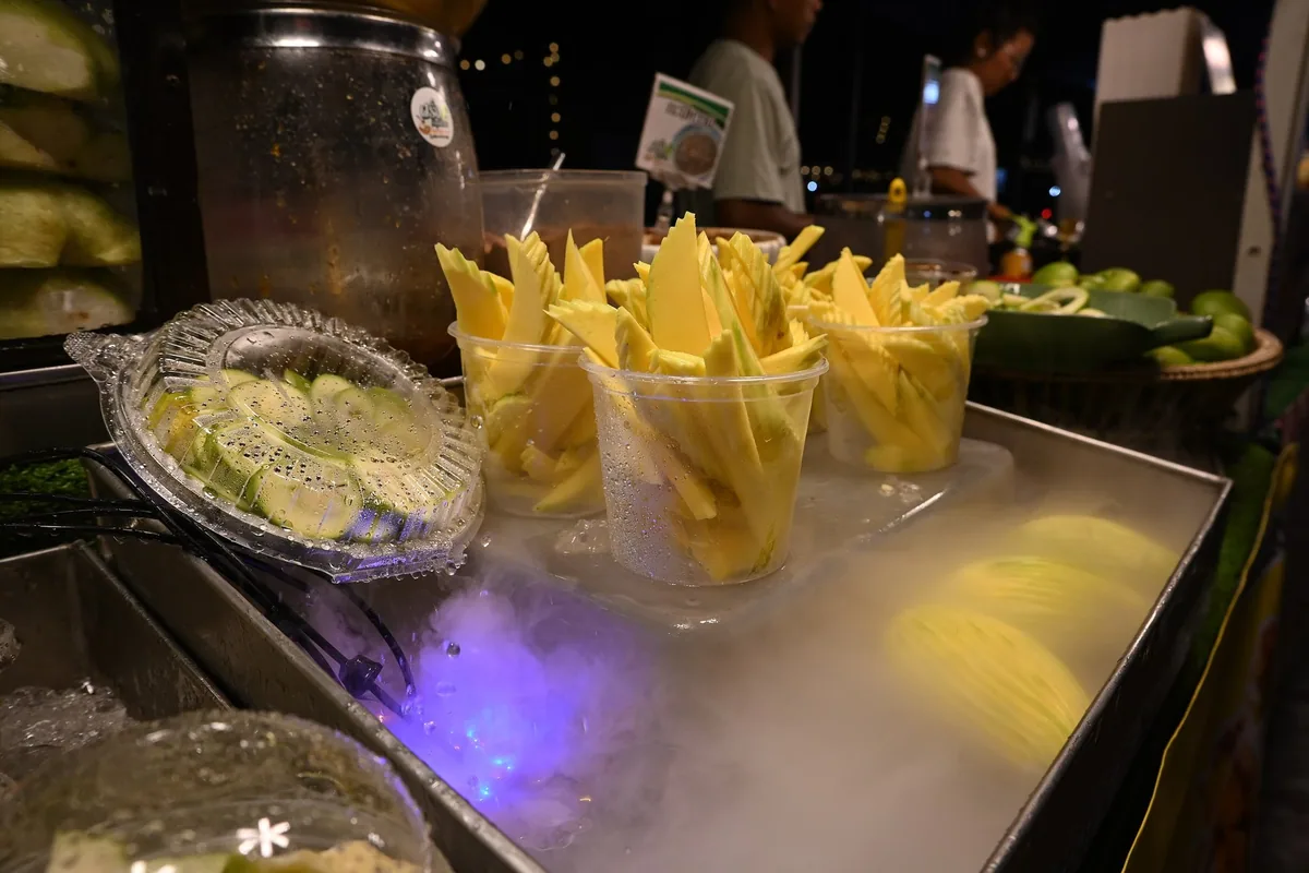 Cups of pre-cut mango and sliced fruit on an iced, misting display tray at a night market stall