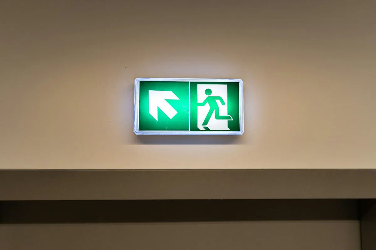 Illuminated green emergency exit sign with a running figure and an arrow, mounted high on a corridor wall