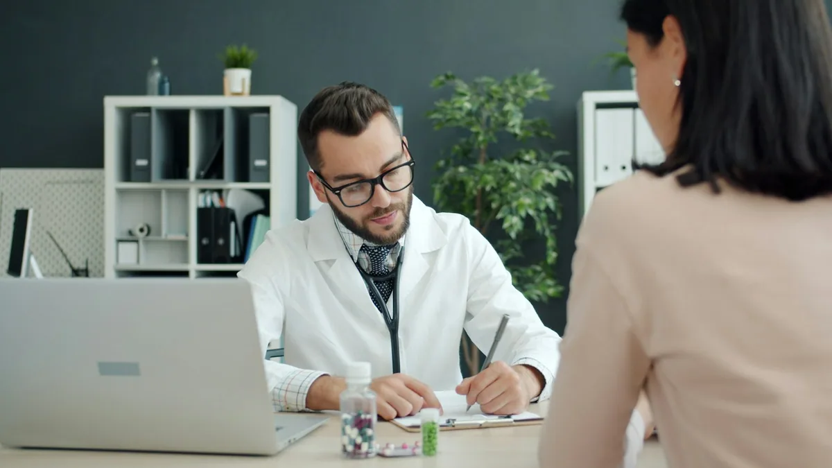 A doctor writing notes at a desk while a patient sits opposite