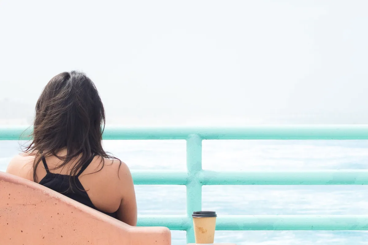 Woman seen from behind sitting alone at a pale turquoise railing, looking out over a hazy sea