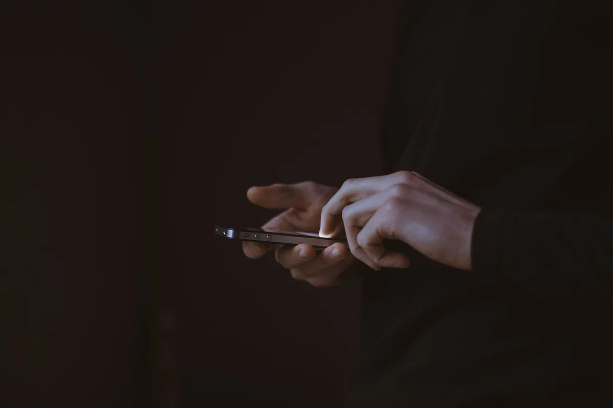 Hands tapping on a smartphone screen in a dark room, the glow lighting the fingertips