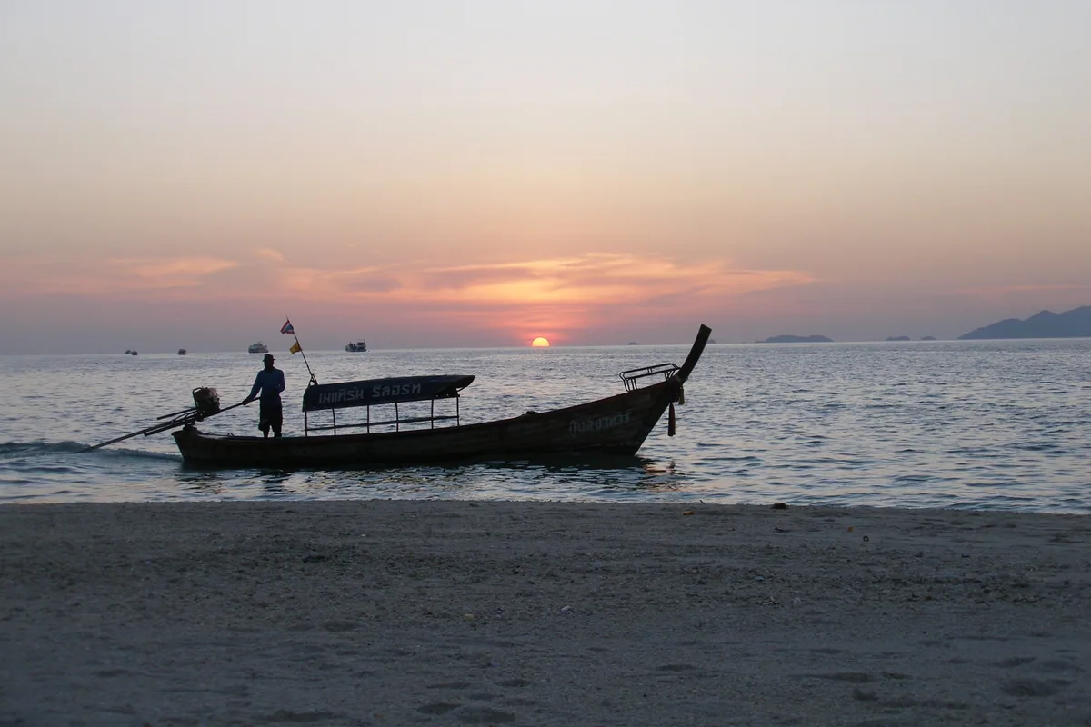 A boatman steering a longtail boat off a Koh Lipe beach at sunset, with islands on the horizon