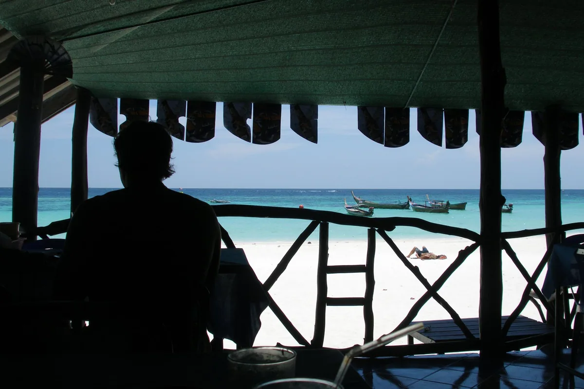 View from a shaded beach shack on Koh Lipe across white sand to longtail boats in the clear shallows