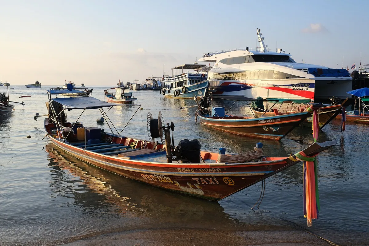 Longtail boats in the shallows beside high-speed ferries moored at Mae Haad pier on Koh Tao