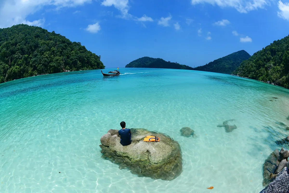 A snorkeler resting on a boulder in clear turquoise shallows of a forested bay in the Surin Islands as a longtail boat passes