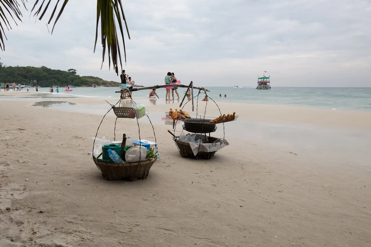 Beach vendor's woven baskets on the white sand of Koh Samet, with swimmers and a boat in the calm bay