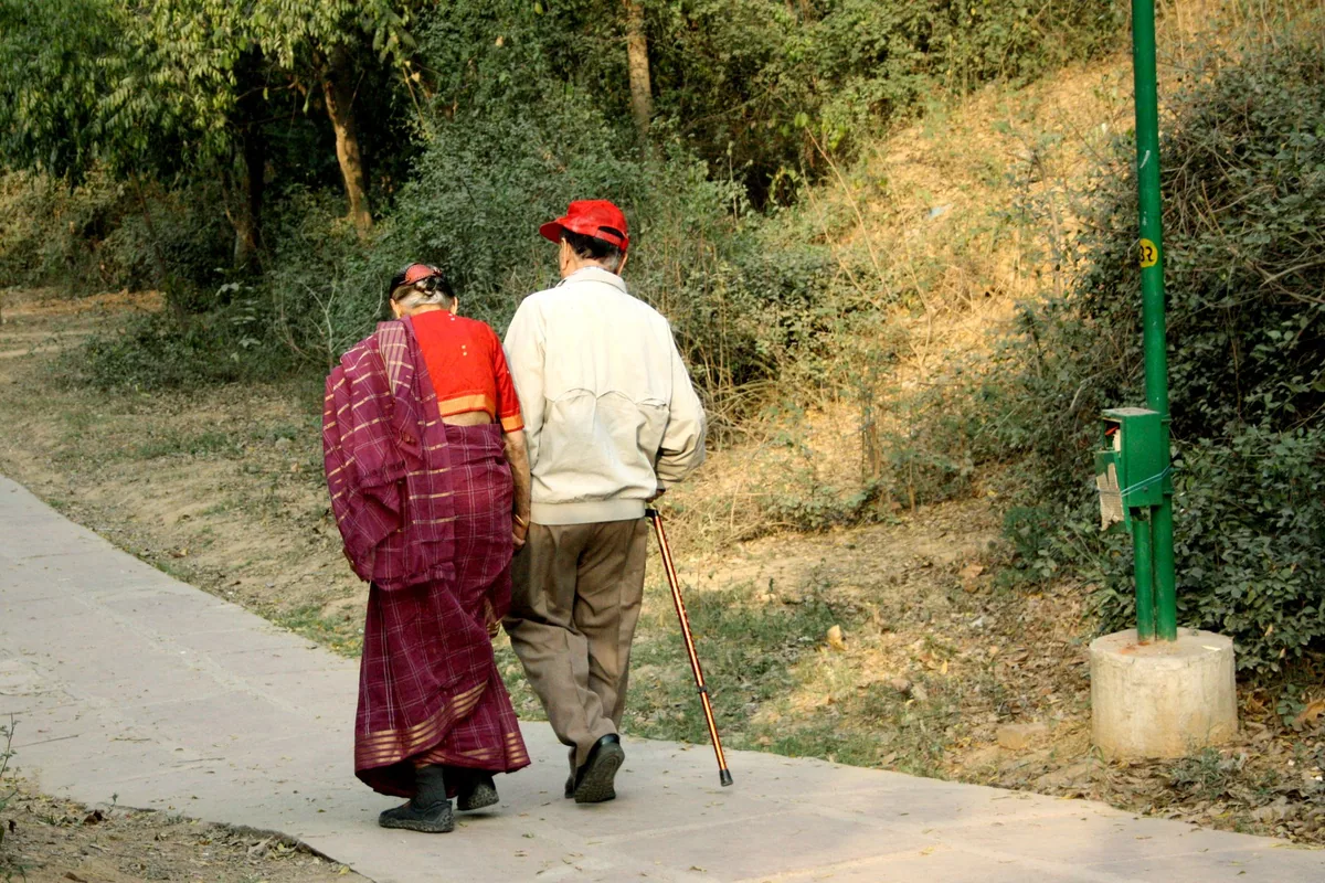 An elderly couple seen from behind walking slowly arm in arm along a park path, the man leaning on a walking cane