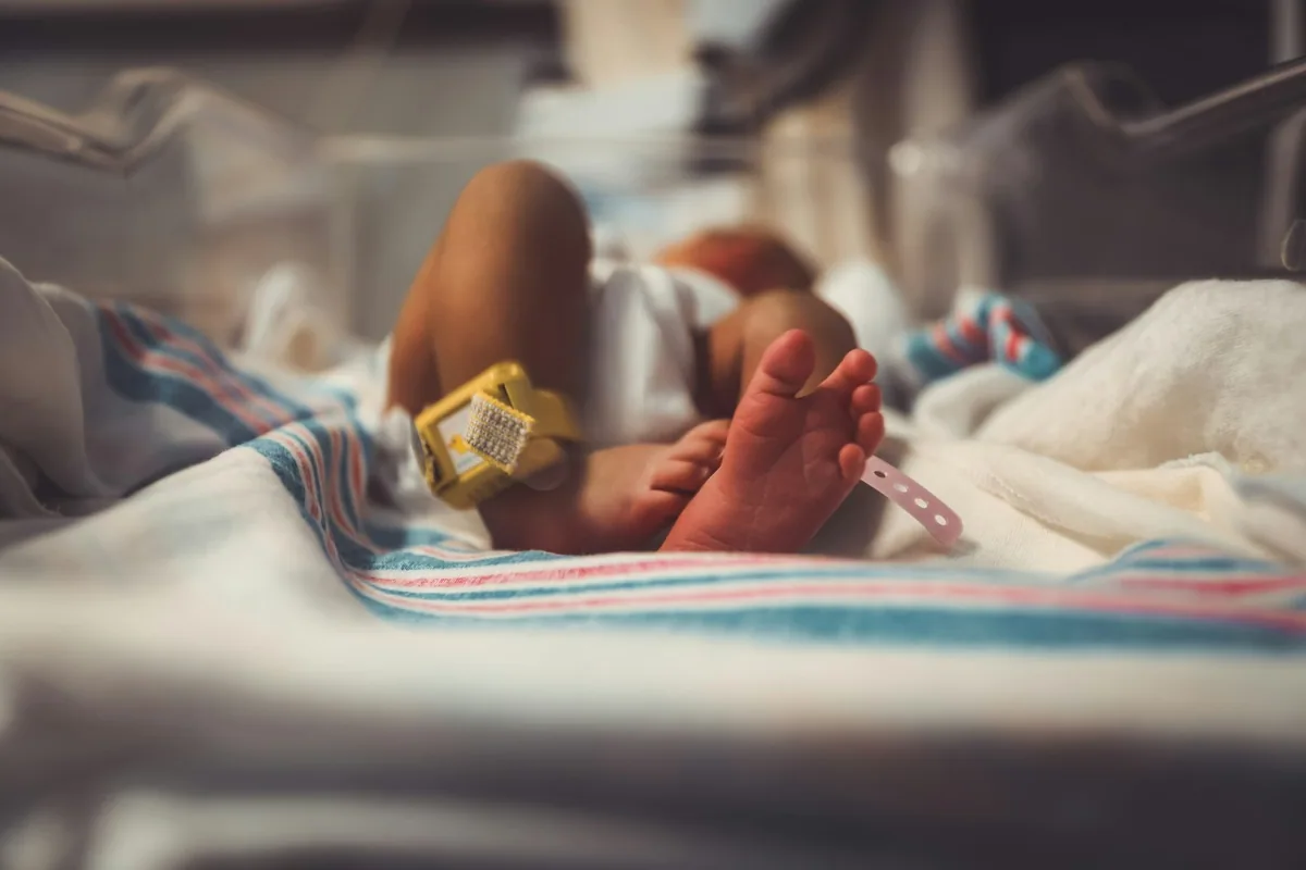 A newborn baby's feet with a hospital name tag and security band, lying on a striped blanket in a hospital cot