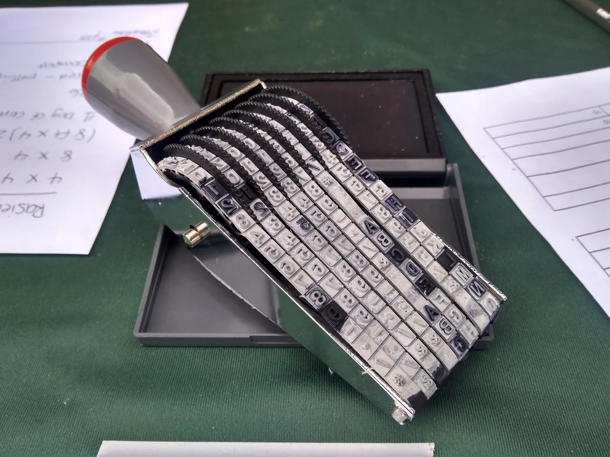 Adjustable rubber stamp with character wheels resting on an open ink pad beside paper forms on a desk