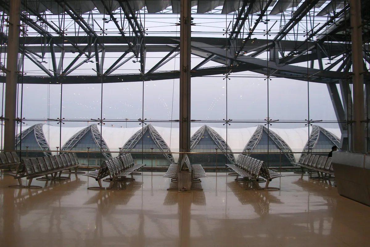 Quiet rows of seats behind the glass wall of Suvarnabhumi Airport, with the terminal's curved roof beyond
