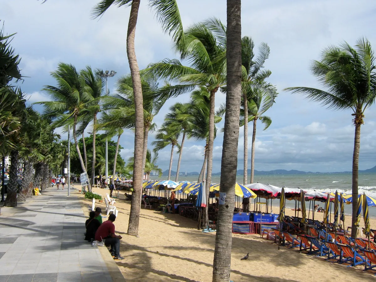 Palm-lined seafront promenade at Jomtien Beach, with people resting on the kerb beside rows of deckchairs and parasols