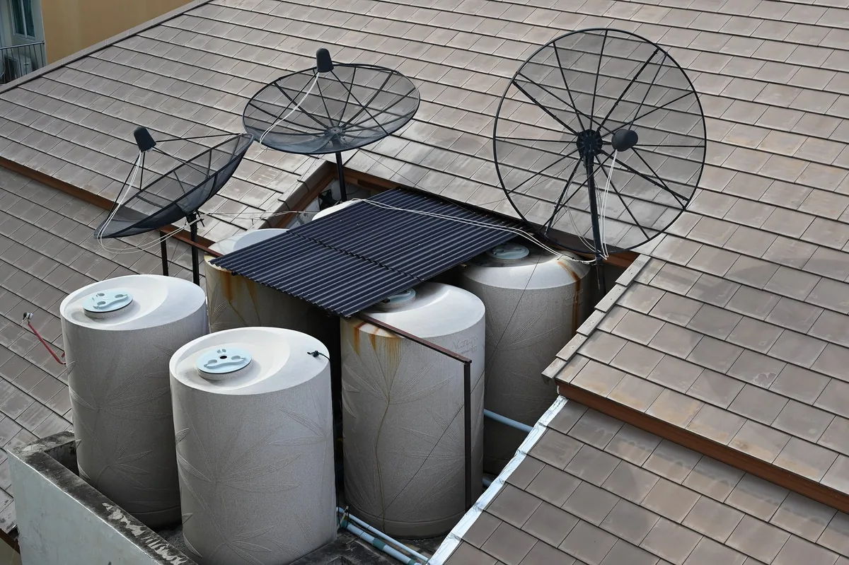 Four white water storage tanks and satellite dishes set into a gap in a tiled roof on a house in Pattaya
