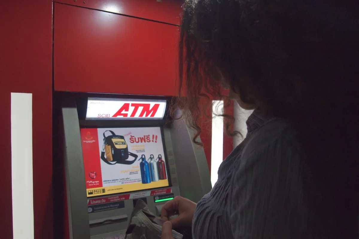 A traveller seen from behind taking cash from a red Siam Commercial Bank ATM on Koh Chang