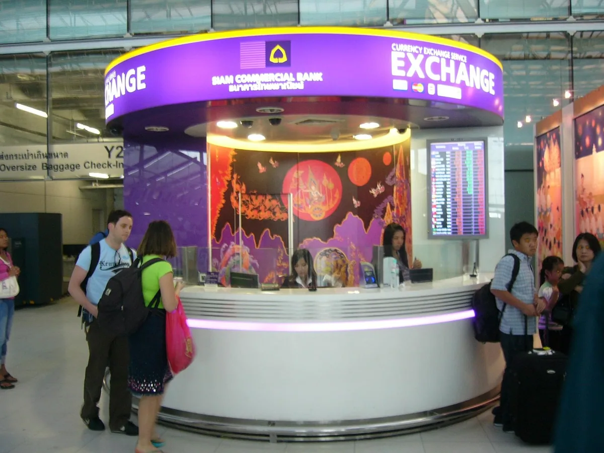 Travellers with backpacks at a round currency exchange counter with a rates board inside Suvarnabhumi Airport