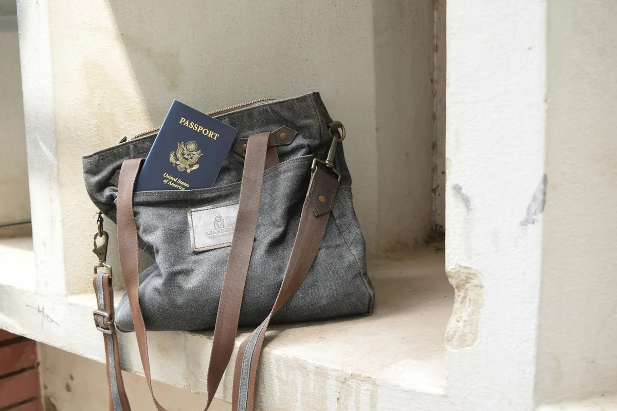 A passport tucked into the outer pocket of a grey canvas shoulder bag resting on a sunlit stone ledge
