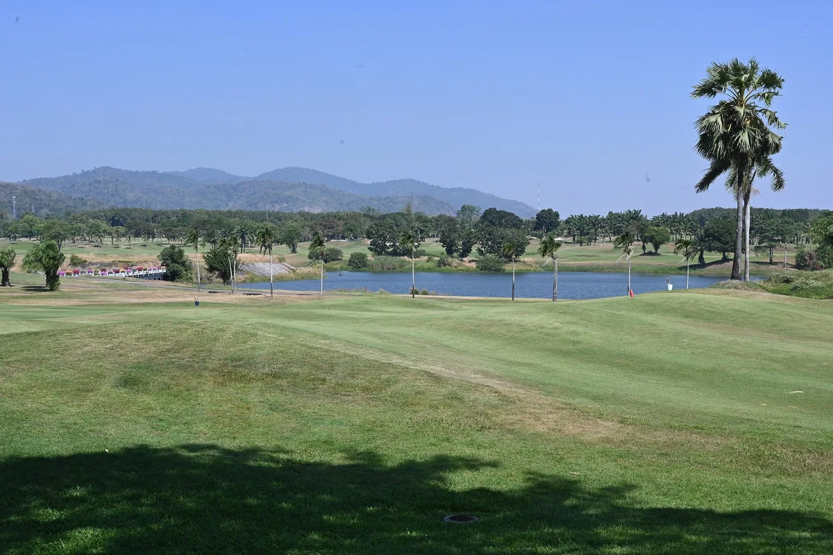 A sunlit fairway sloping to a lake and a tall palm, with forested hills behind, on a golf course in Chonburi province