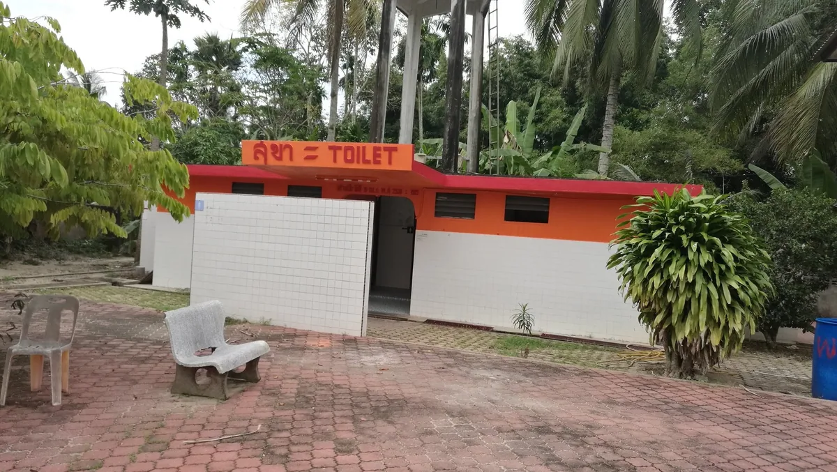 A small orange and white public toilet block with a bilingual Thai and English TOILET sign among palm trees