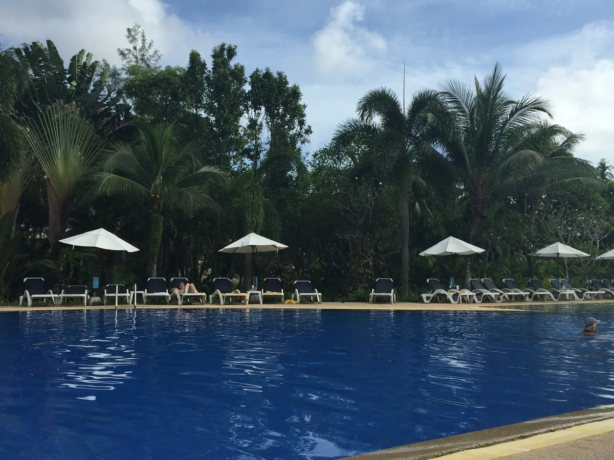 Sun loungers and white parasols lining a large resort pool under palm trees at Karon Beach, Phuket