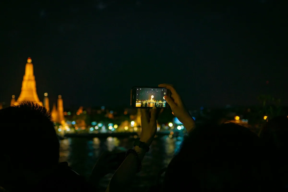 A visitor holding up a smartphone to photograph the floodlit Wat Arun across the Chao Phraya River at night