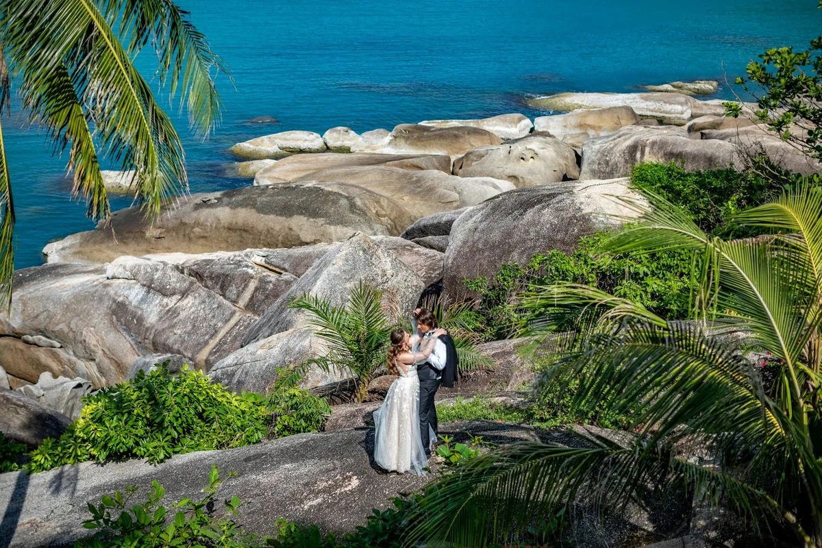 Newlyweds embracing among granite boulders and palms above the turquoise sea on the coast of Koh Samui