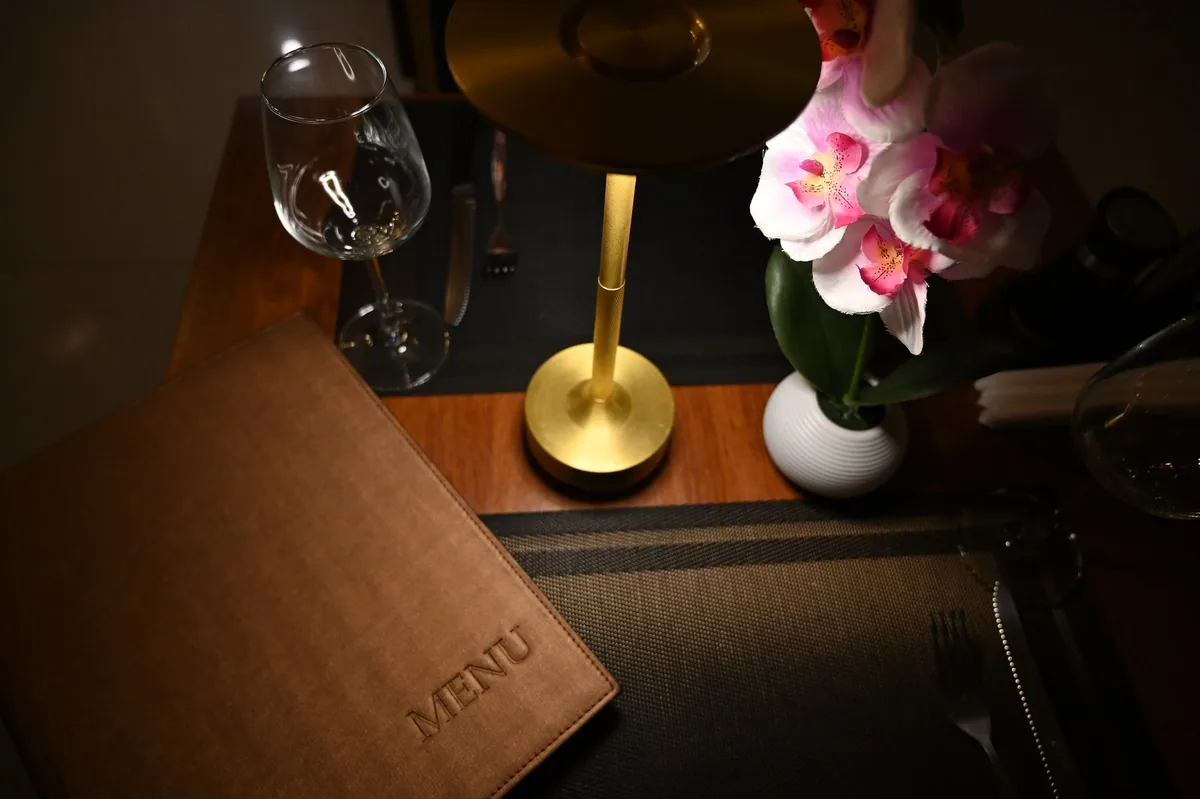 A leather-bound menu on a set restaurant table beside a wine glass, brass lamp and pink orchids, Pattaya