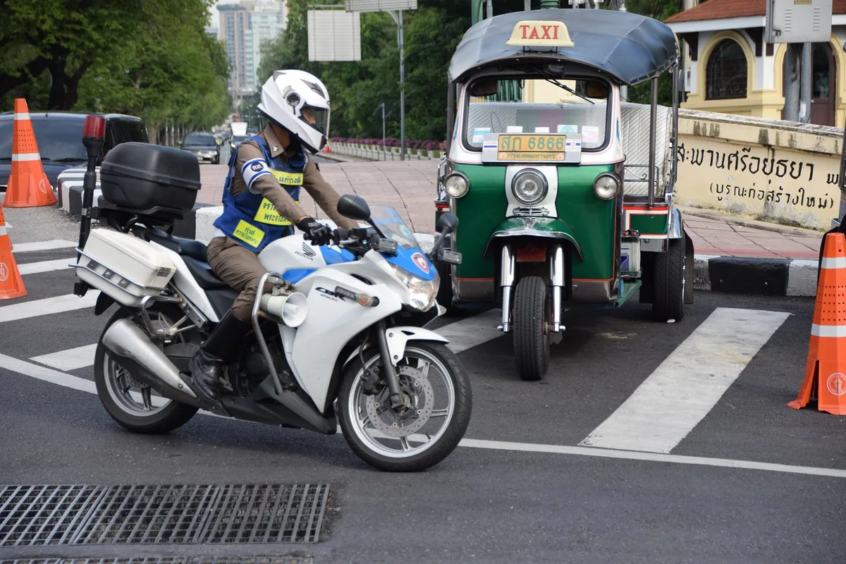 A Thai traffic police officer on a white motorbike beside a green tuk-tuk at a road junction